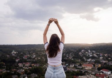 Genç bir kadın, elleri bir kazanan gibi havada bir dağda oturuyor. Görülen manzaradan bir özgürlük ve memnuniyet jesti. Yukarıdan güzel bir şehir manzarası. Tatil, dağlarda yürüyüş, aktif yaşam tarzı.