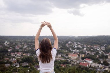 Genç bir kadın, elleri bir kazanan gibi havada bir dağda oturuyor. Görülen manzaradan bir özgürlük ve memnuniyet jesti. Yukarıdan güzel bir şehir manzarası. Tatil, dağlarda yürüyüş, aktif yaşam tarzı.