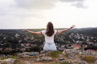 Genç bir kadın, elleri bir kazanan gibi havada bir dağda oturuyor. Görülen manzaradan bir özgürlük ve memnuniyet jesti. Yukarıdan güzel bir şehir manzarası. Tatil, dağlarda yürüyüş, aktif yaşam tarzı.
