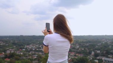 Genç bir kadın telefonuyla dağın tepesinden şehrin güzel bir manzarasının fotoğrafını çekiyor. Akıllı telefondan fotoğraflar ve videolar. Seyahat blogcusu ve etkileyicisi