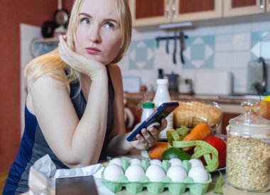 Bir kadın akıllı telefon uygulamasında yemeğin kalori miktarını kontrol eder. Kilo kaybı için kalori sayılıyor. Masada diyet için ürünler var.