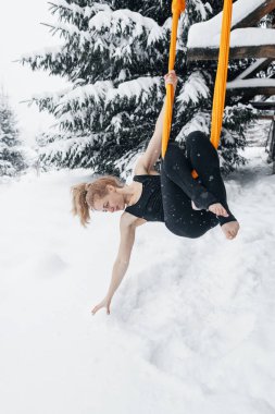Dağlardaki karlı ormanda sinek yogası yapan genç bir kadın. Kışın hamakta Yoga, kar ve köknar ağaçlarına karşı. Olağanüstü koşullarda jimnastik numaraları.