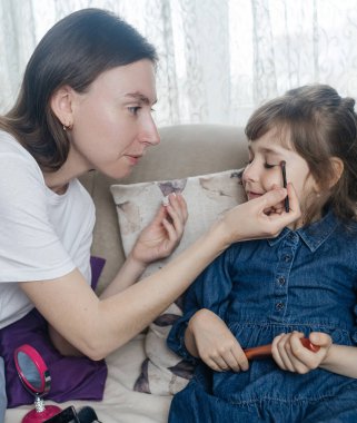 Anne kızının yüzüne makyaj yapıyor ve birlikte takılıyorlar. Ailedeki kadınlar arasındaki iletişim. Eğlence zamanı. Mutlu aile kavramı. Küçük kız makyajını yapıyor.