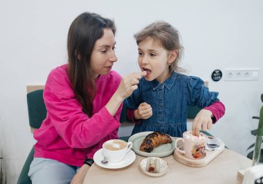 Anne ve kızı bir kafede, kahve ve kakao içiyor, kruvasan ve kek yiyor. Eğleniyoruz ve birlikte vakit geçiriyoruz.
