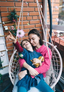 Anne ve kızı, çiçeklerle çevrili bir hasır bahçede salıncakta sarılıyorlar. Mutlu bir çocukluk ve sevgi dolu bir aile kavramı.