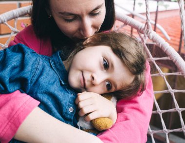 Anne ve kızı, çiçeklerle çevrili bir hasır bahçede salıncakta sarılıyorlar. Mutlu bir çocukluk ve sevgi dolu bir aile kavramı.