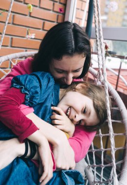 Anne ve kızı, çiçeklerle çevrili bir hasır bahçede salıncakta sarılıyorlar. Mutlu bir çocukluk ve sevgi dolu bir aile kavramı.
