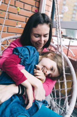Anne ve kızı, çiçeklerle çevrili bir hasır bahçede salıncakta sarılıyorlar. Mutlu bir çocukluk ve sevgi dolu bir aile kavramı.