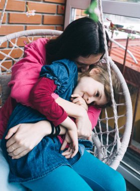 Anne ve kızı, çiçeklerle çevrili bir hasır bahçede salıncakta sarılıyorlar. Mutlu bir çocukluk ve sevgi dolu bir aile kavramı.