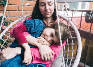 Anne ve kızı, çiçeklerle çevrili bir hasır bahçede salıncakta sarılıyorlar. Mutlu bir çocukluk ve sevgi dolu bir aile kavramı.