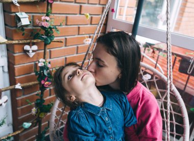Anne ve kızı, çiçeklerle çevrili bir hasır bahçede salıncakta sarılıyorlar. Mutlu bir çocukluk ve sevgi dolu bir aile kavramı.