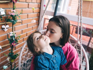 Anne ve kızı, çiçeklerle çevrili bir hasır bahçede salıncakta sarılıyorlar. Mutlu bir çocukluk ve sevgi dolu bir aile kavramı.