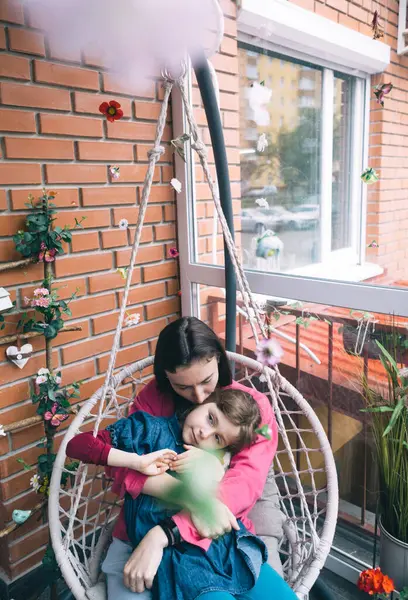 Anne ve kızı, çiçeklerle çevrili bir hasır bahçede salıncakta sarılıyorlar. Mutlu bir çocukluk ve sevgi dolu bir aile kavramı.