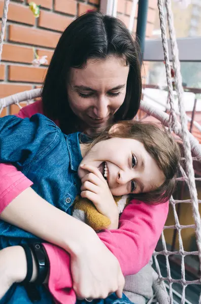 Anne ve kızı, çiçeklerle çevrili bir hasır bahçede salıncakta sarılıyorlar. Mutlu bir çocukluk ve sevgi dolu bir aile kavramı.