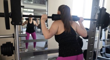 Ayrıca Smith Machine üzerinde egzersiz yapan cüsseli bir kadın. Halterle simülatöre çömelmiş. Kilo kaybı için egzersiz