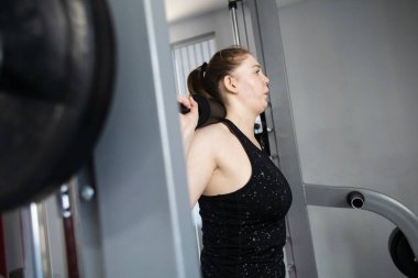 Ayrıca Smith Machine üzerinde egzersiz yapan cüsseli bir kadın. Halterle simülatöre çömelmiş. Kilo kaybı için egzersiz