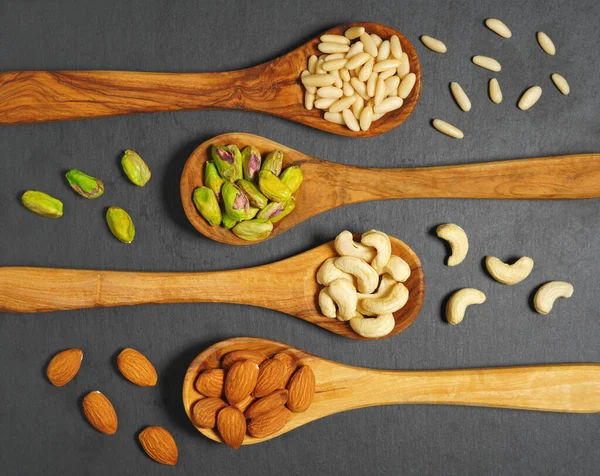 Nuts mix with pine nuts, pistachios, cashews nuts and almonds in wooden spoons top view closeup on dark gray background.