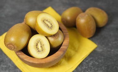 Gri arka planda dilimlenmiş kivi ile ahşap bir kasede altın sarısı bir meyve..