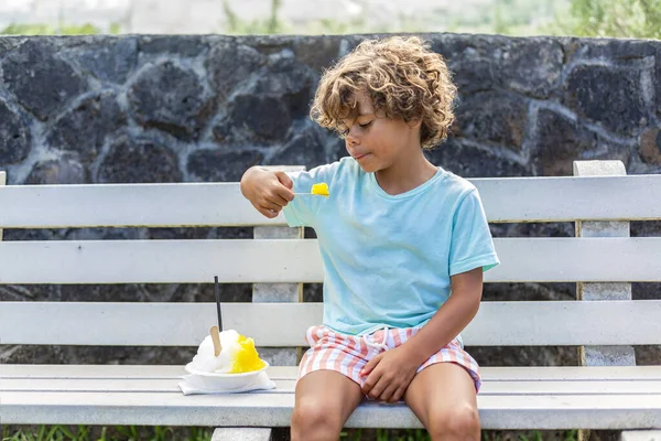 スノーコーンを食べたり 公共の通りのベンチで屋外で氷を剃ったりするか かわいい多様な少年 暑い夏の日に涼しいリフレッシュを楽しむ ストック画像