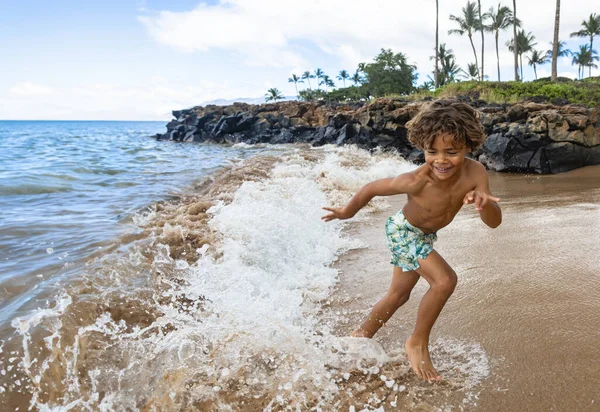 家族の休暇中に若いアフリカ系アメリカ人の少年がビーチで走ったり遊んだりして笑っています 海の波の中で遊ぶのは楽しいし活発なので ロイヤリティフリーのストック画像