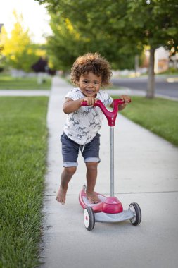 Şirin, gülümseyen, çeşitli genç bir çocuk mahallesinde eğlenceli bir yaz gününde kaldırımda scooterını sürüyor. Farklı çocuklar için yaşam tarzı fotoğrafı. Güvenli Mahalle konsepti fotoğrafı