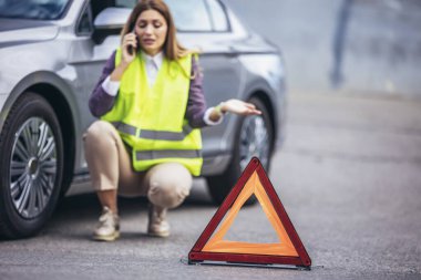 Genç beyaz kadın, arabası yolda bozulmuşken cep telefonuyla konuşuyor. Yansıtıcı uyarı üçgenleri var. .