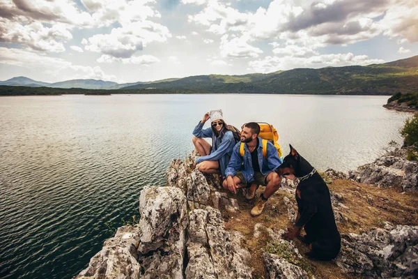 山の湖で楽しむ崖の上に座っているバックパックと彼らのDobermanを持つハイカー — ストック写真