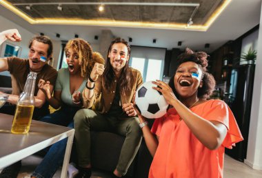 Young group watching sports on television and cheering. Emotional football fans watching football game together at home. Sports Fans with Painted Faces Sitting on a Couch Watch Game on TV
