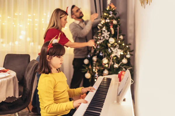 圣诞节期间 年轻的父母装饰圣诞树 欣赏女儿弹钢琴时的音乐 — 图库照片