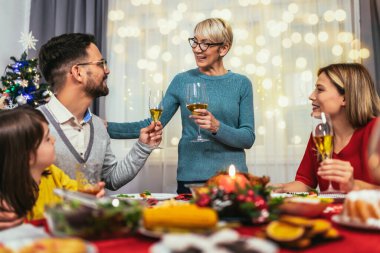 Fotoğraf, sevgili bir büyükanne, anne, baba ve kızıyla bir Noel yemeğinin sıcaklığını ve neşesini yansıtır. Aile şenlik masasının etrafında toplanmış, gülümsüyor ve gülüyor. Geleneksel hindi ziyafeti, hindi dolması ve diğer şeyler gibi.