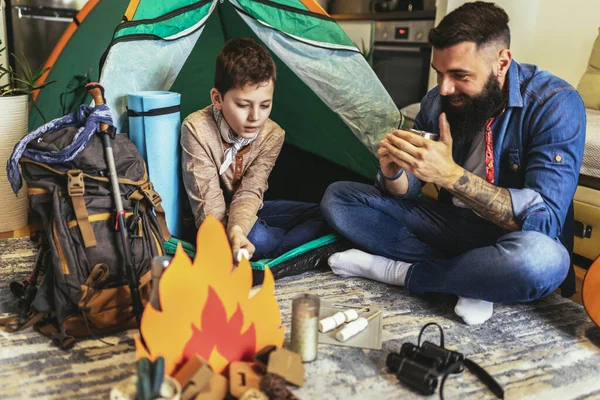 Dad with his son camp inside their home. They have pitched a tent and have a fake campfire.