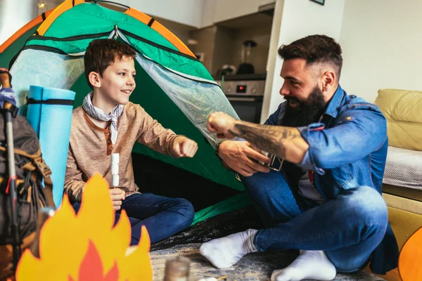Dad with his son camp inside their home. They have pitched a tent and have a fake campfire.