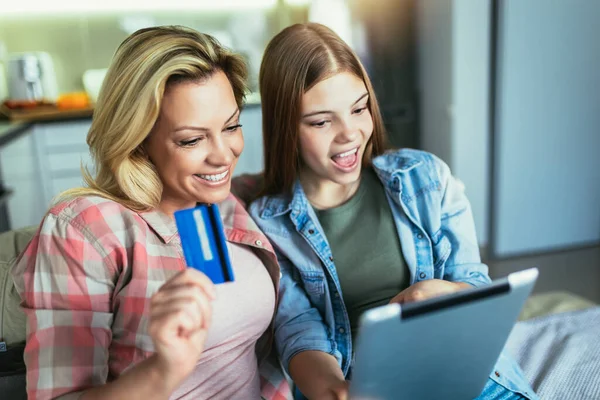 Mother and daughter shopping together on digital tablet, holding credit card, sitting and hugging on couch at home.