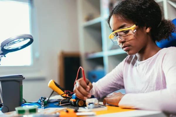 Smart Young African American Schoolgirl is Studying Electronics and Soldering Wires and Circuit Boards in Her Science Hobby Robotics Project. Girl is Working on a Robot in Her Room. Education Concept.