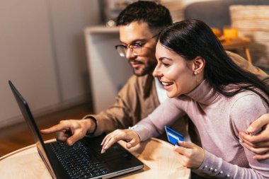 Happy young couple shopping together, customers paying by credit card online, sitting in cozy living room at home