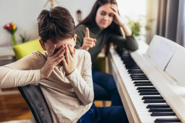 Brother and sister play electric piano at home. The sister helps her younger brother to play piano. The boy is angry that he has to play piano.