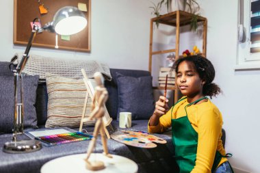 Black preteen girl happily at a table, surrounded by an array of creative materials for painting to inspire their next great idea.