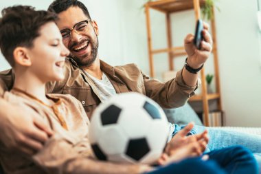 Boy watching soccer match with father at home, sitting on sofa