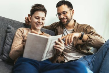 Happy boy and his cheerful dad are reading a book sitting on the couch at home in the living room.