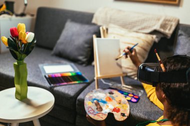 Evde boya fırçasıyla resim yapan mutlu siyah liseli kız kendini meta-evrene daldırmak için VR gözlükleri kullanıyor.