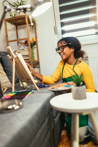 Black preteen girl happily at a table, surrounded by an array of creative materials for painting to inspire their next great idea.