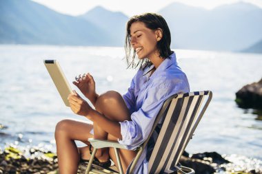 Güneşli bir günde deniz kenarındaki iskemlede dijital tablet kullanan gülümseyen kadın.