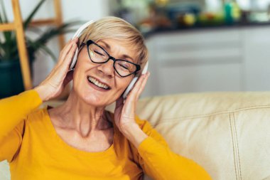 Evde müzik dinleyen, kulaklıklı, mutlu, yaşlı bir kadın.
