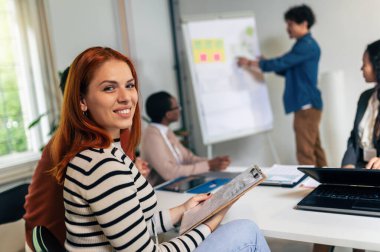 Genç bir kadın, takım lideri bir grup genç meslektaşına sunum yaparken kameraya bakıyor.