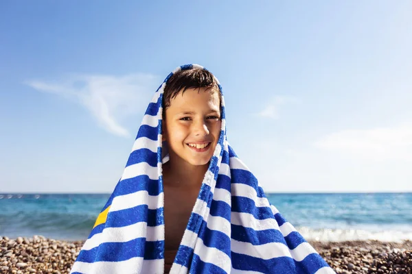海滩上的小男孩头戴一条蓝白相间的条纹浴巾 开心地笑着 — 图库照片