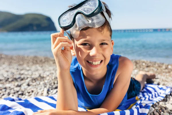 穿着潜水护目镜的小男孩躺在一条蓝白相间的沙滩毛巾上 开心地笑着 — 图库照片