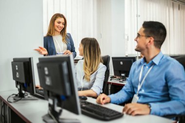 Bir grup öğrenci bilgisayar laboratuvarında bir masanın önünde oturuyorlar. Her biri ekranlarına odaklanmış durumda. Bayan öğretmenleri her öğrenciyi tek tek kontrol etmek için etrafta dolanıyor..