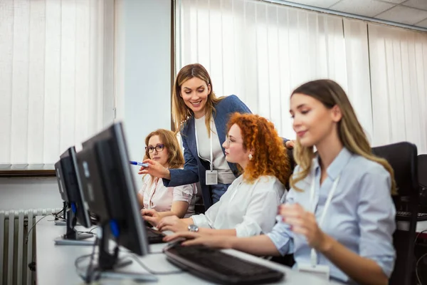 学生のグループは コンピュータ研究室のデスクトップの前に座っています 各生徒を個別にチェックするために女性教師が彼女の方法を作るように 彼らはそれぞれの画面に焦点を当てています — ストック写真