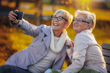 İki yaşlı kadın selfie çekiyor ve gülümsüyor. Sonbaharda bir bankta oturuyorlar.