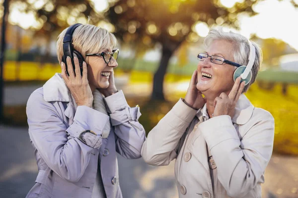Two older women listening to music on their wireless headphones in the colorful park in the autumn